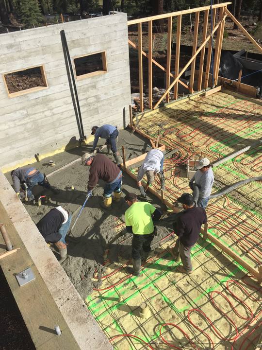 Gonzalez Concrete's crew placing and screeding a slab over radiant-heat tubing and wire mesh, inside board-formed foundation walls with the house framing already going up