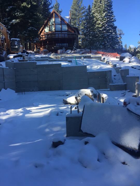 Poured foundation walls and stem walls under fresh snow, with an excavator on site and a finished log home behind