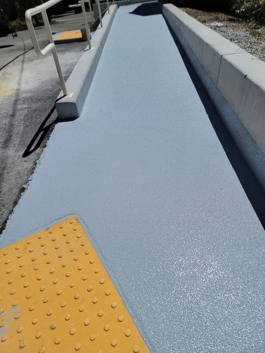 A resurfaced concrete access ramp coated in grey non-slip finish, with a yellow truncated-dome warning pad at the base