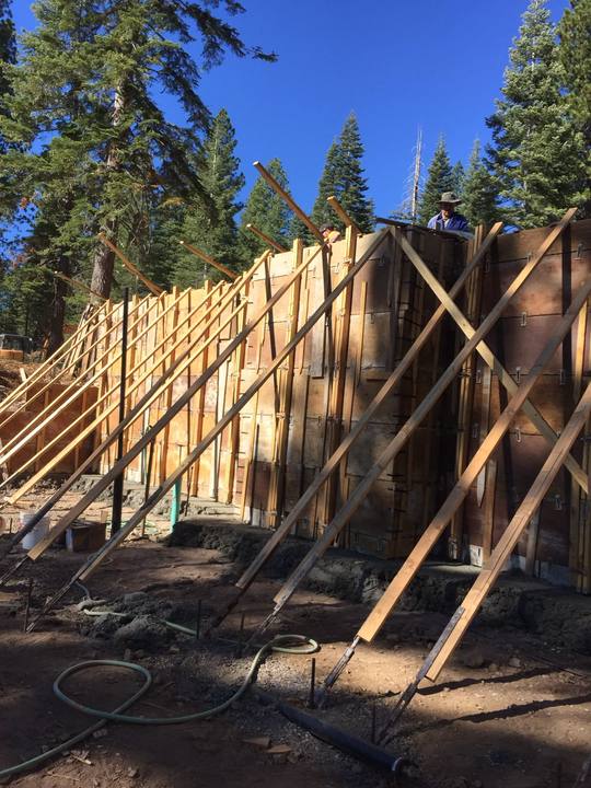 A tall foundation wall in braced plywood forms, kickers running down to stakes, with two of the crew working from the top of the wall