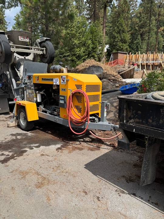 A Putzmeister line pump hooked to a mixer truck at the top of a driveway, hose running downhill to formed foundation walls in the trees