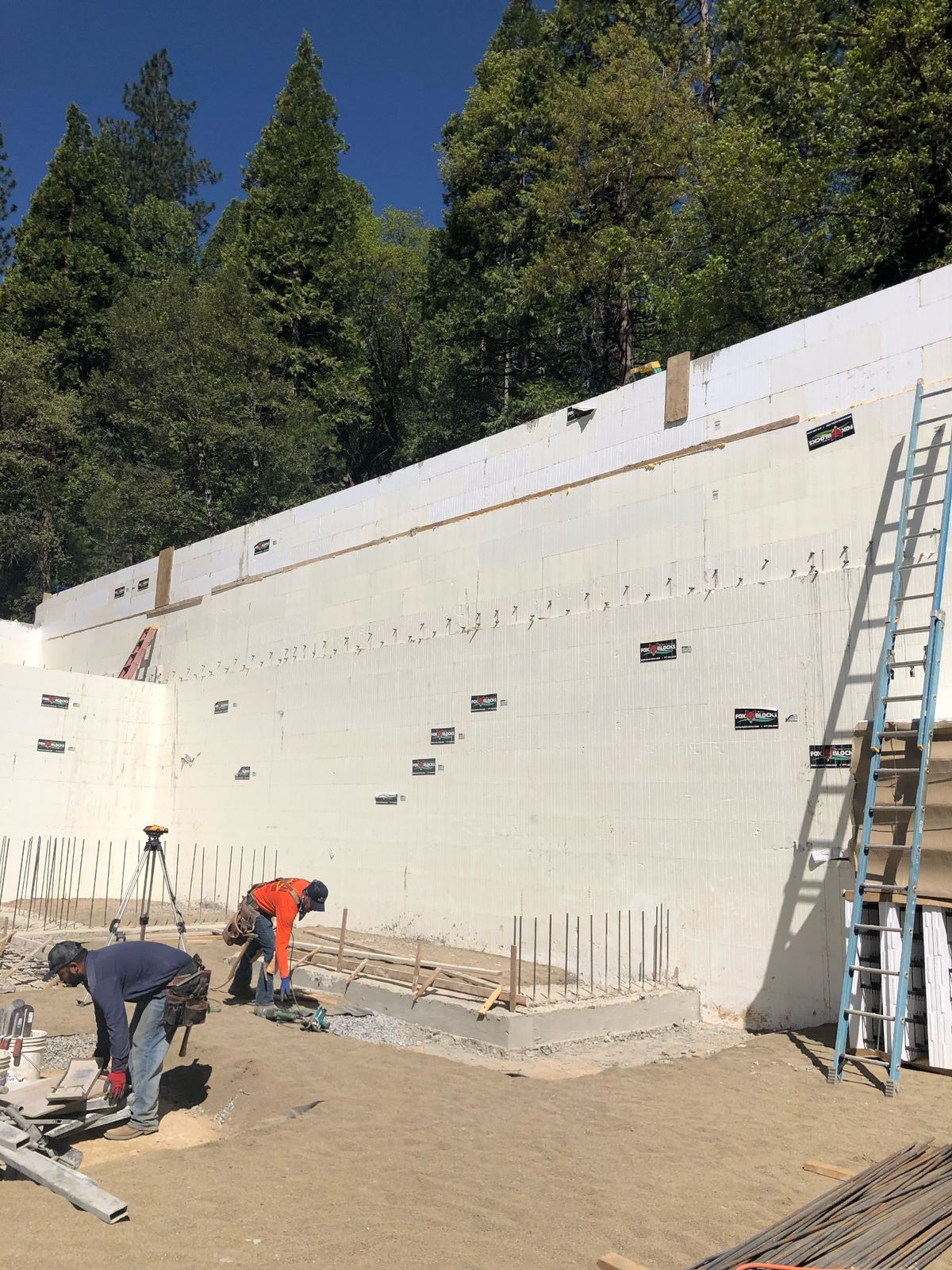 An insulated concrete form (ICF) wall stacked full height on a poured footing against a cut slope, with the crew shooting grades off a survey level