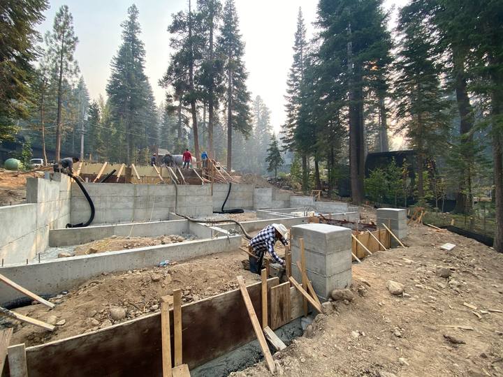 Poured stem walls and column pads across a large foundation footprint, forms still standing and the crew working the far wall