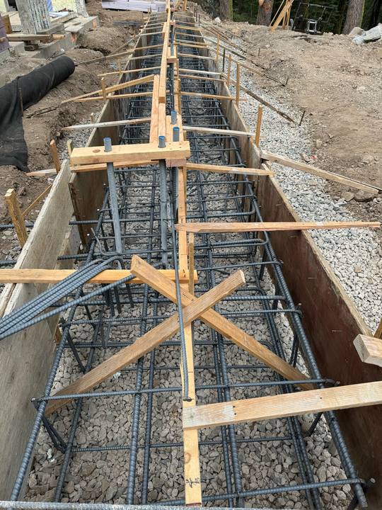 A deep footing trench with a heavy rebar cage tied inside it and hold-down bolts hung off the top, formed and ready for concrete